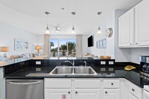 Une cuisine moderne avec un plan de travail noir, un évier en acier inoxydable et des meubles blancs. On y trouve des luminaires suspendus, un ventilateur de plafond et une vue sur les palmiers à l’extérieur.
