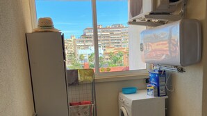 Une petite buanderie comprenant un lave-linge, un sèche-linge, une fenêtre donnant sur des immeubles, un ventilateur de plafond et un climatiseur fixé au mur.
