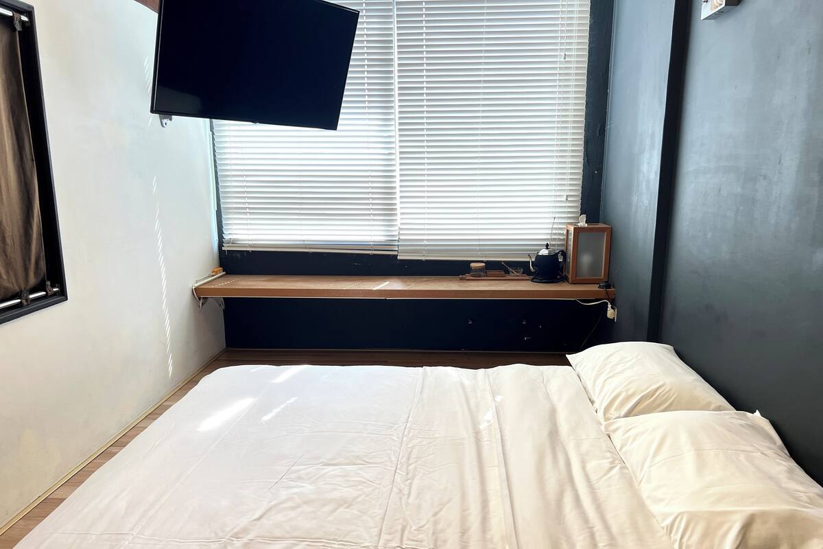Ein Schlafzimmer mit einem Bett, einem an der Wand montierten Fernseher, einem Fenster mit Jalousien und einem kleinen Regal mit Wasserkocher und Bilderrahmen.