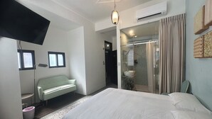 A hotel room with a large bed, a sitting area with a bench, a TV mounted on the wall, a shower area with a glass door, and a ceiling fan. - Hour Roots (Tainan)