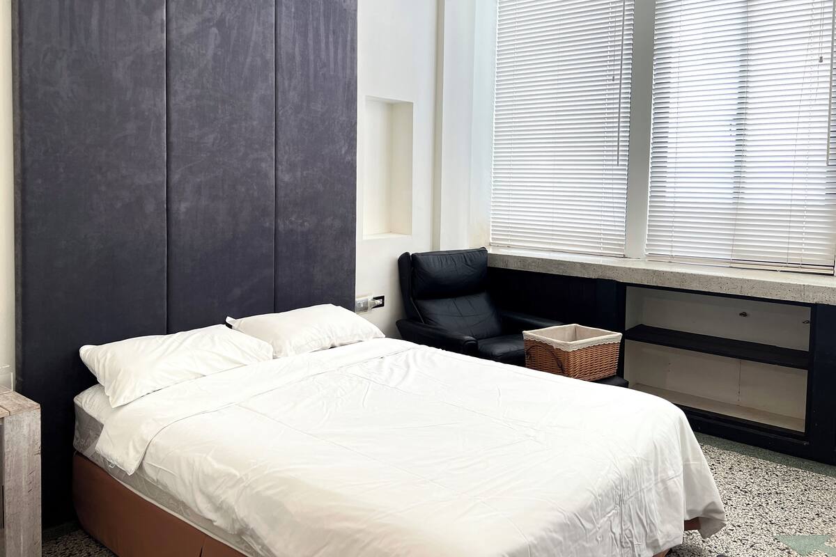 Ein Schlafzimmer mit einem Bett, einem dunklen Kopfteil, einem Fenster mit Jalousien, einem schwarzen Sessel und einem kleinen Korb.