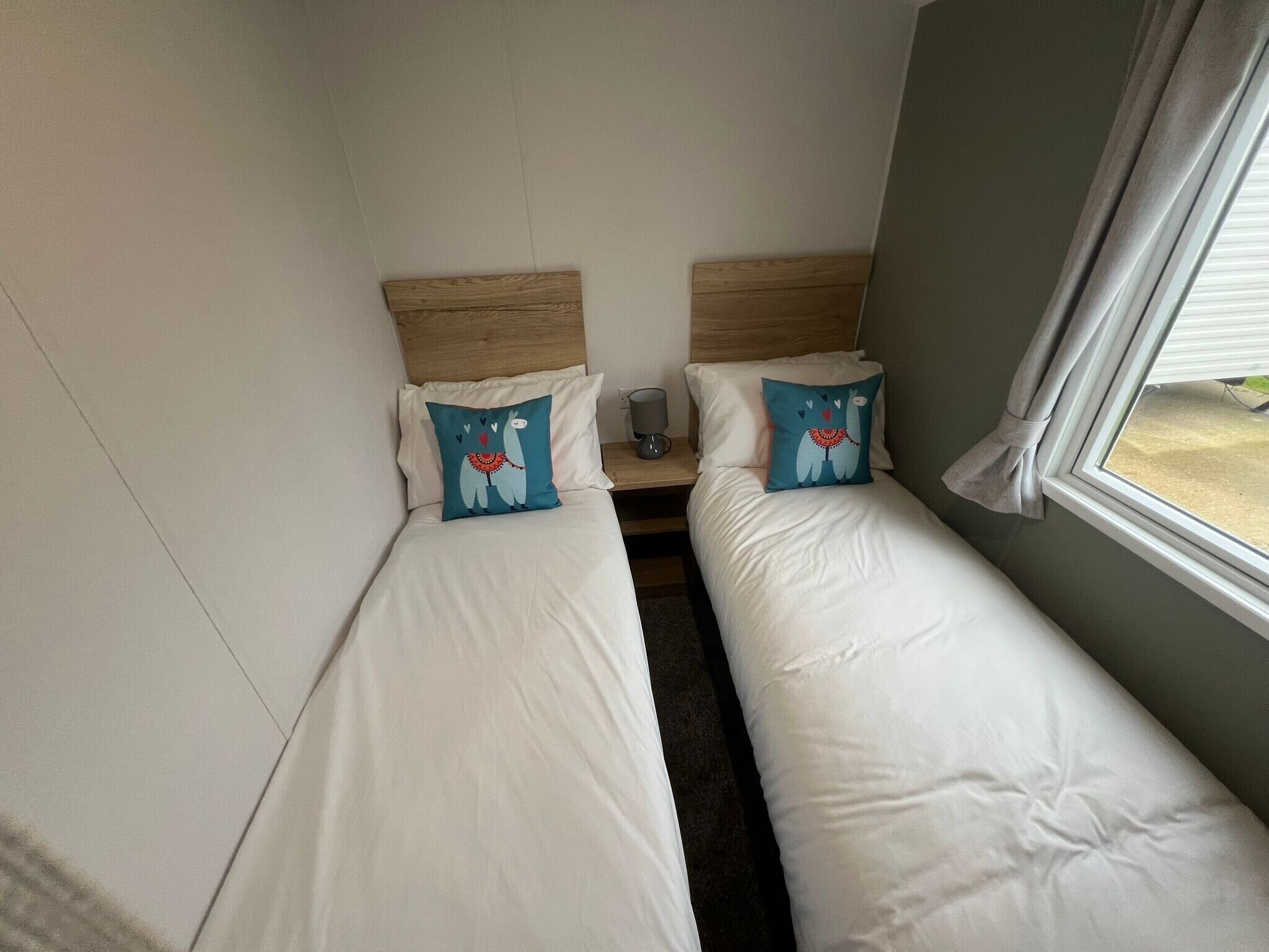 A small, cozy room with two single beds, each with a matching pillow and a wooden headboard. A small table with a lamp is between the beds.