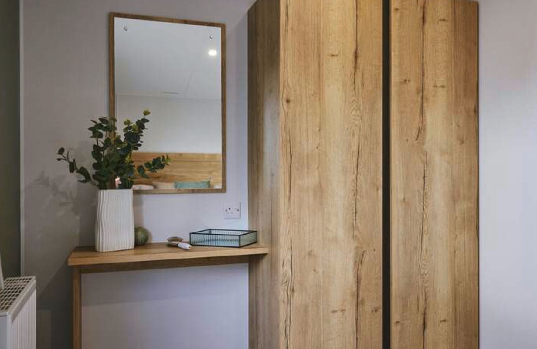 Ein Zimmer mit einem Holzschrank, einem weißen Heizkörper, einem kleinen Tisch mit Topfpflanze und einem Spiegel an der Wand.