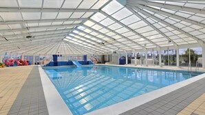 An indoor swimming pool with a high ceiling and glass panels, surrounded by tiled flooring and a clear view of the outside.