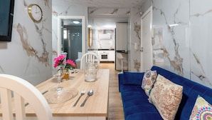 A modern living room with a blue sofa, a dining table set with cutlery, and a kitchen in the background.
