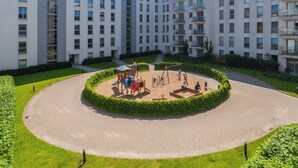 Un parque infantil rodeado de una zona pavimentada con diseño circular, con edificios de apartamentos modernos al fondo.