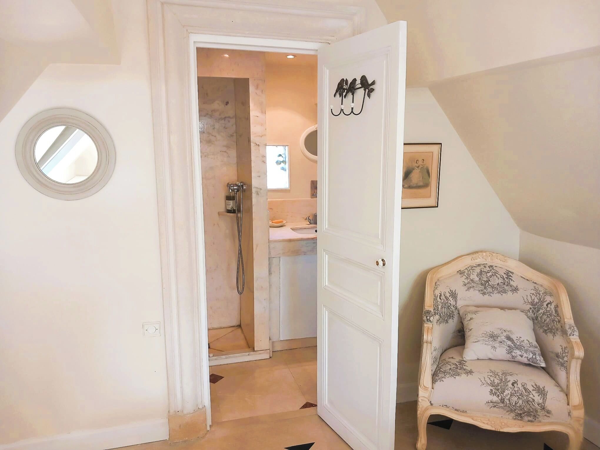 A room with a door leading to a bathroom, a round mirror, a wooden chair with floral upholstery, and a framed picture on the wall.