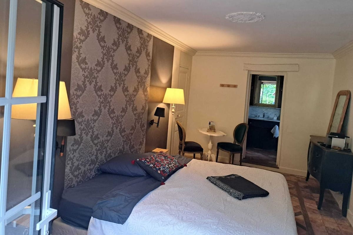 A hotel room with a bed, two bedside lamps, a small table with a lamp, a chair, a mirror, and a view of the bathroom.