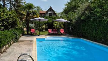A swimming pool with a metal ladder, surrounded by lush greenery and red lounge chairs.