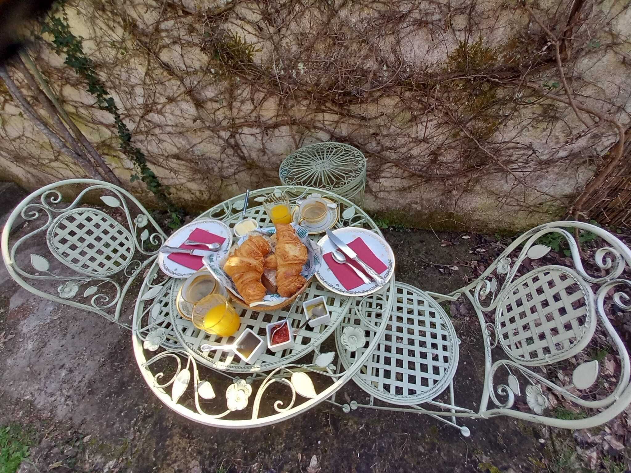 Une table en métal placée à l’extérieur, sur laquelle se trouvent un panier de croissants, un verre de jus d’orange et une tasse de café.