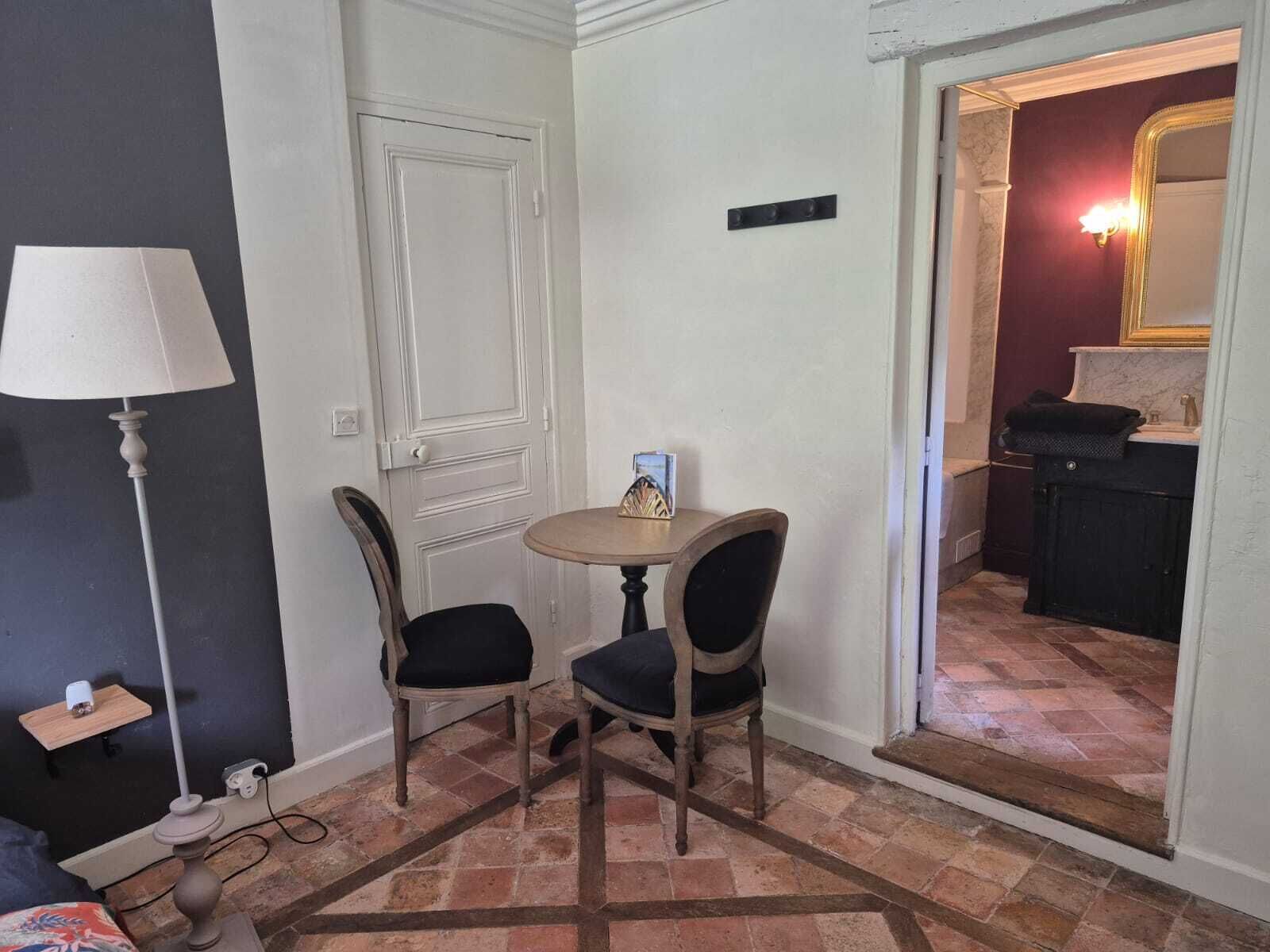 Une petite pièce avec une porte blanche, une table ronde avec deux chaises, une lampe et un miroir dans un coin.