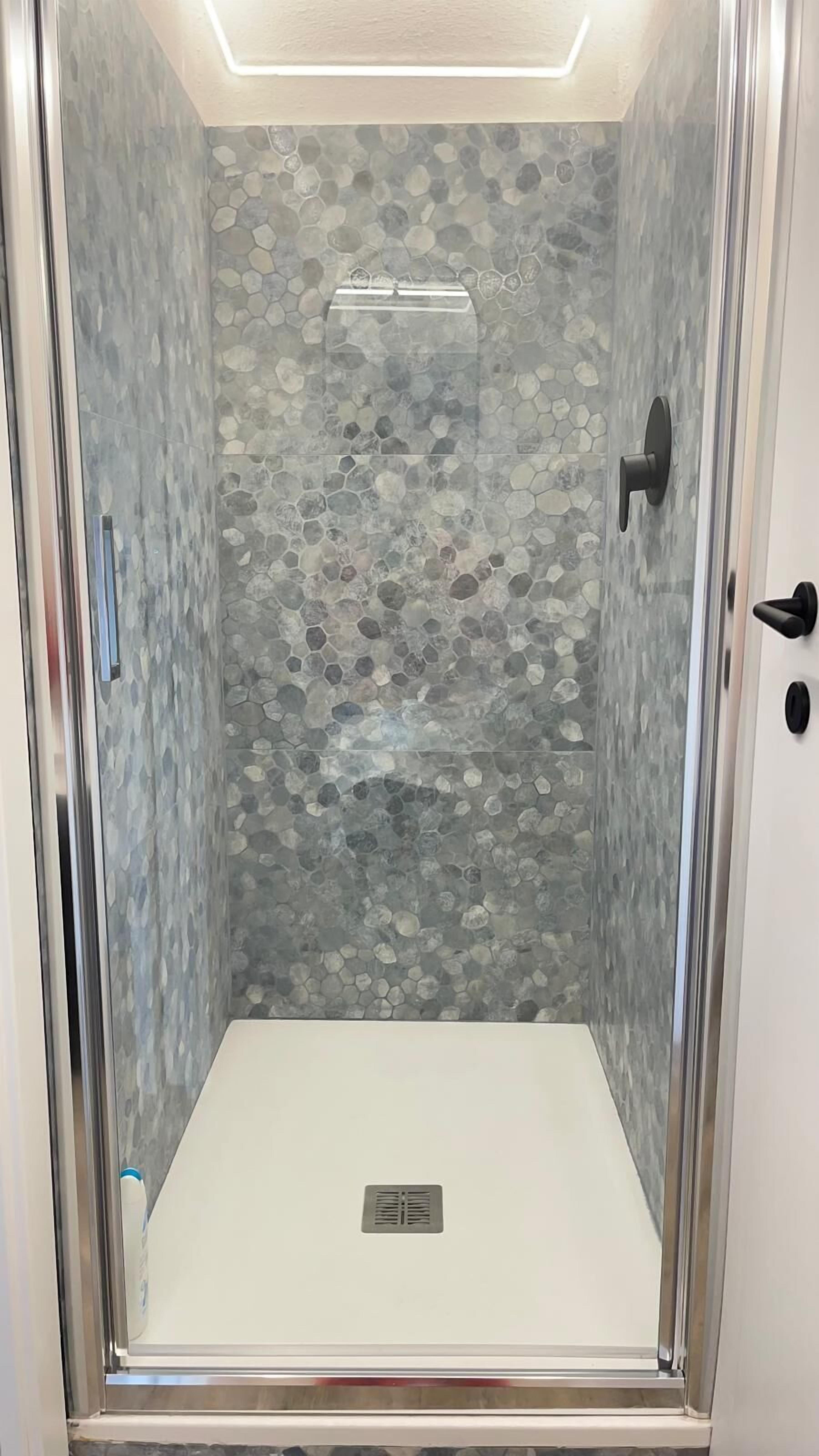 A glass-enclosed shower with a stone mosaic wall, a black door handle, and a ventilation grate.
