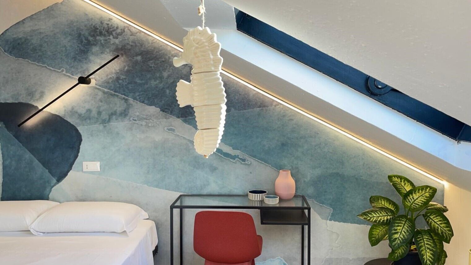A modern room with a bed, a red chair, a glass table, a potted plant, and a seahorse decoration.