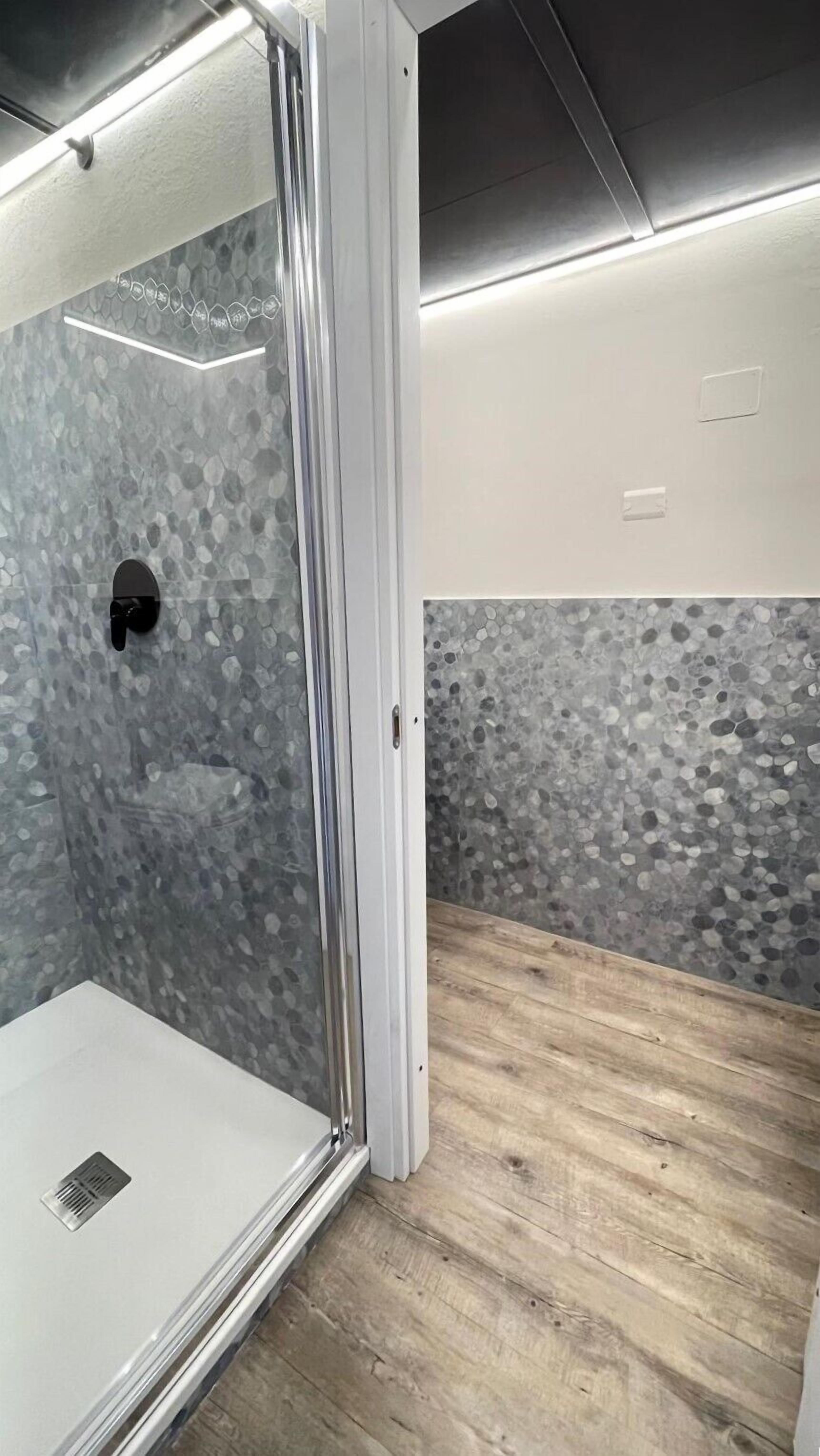 A modern bathroom with a glass shower enclosure, pebble-patterned walls, and wooden flooring.
