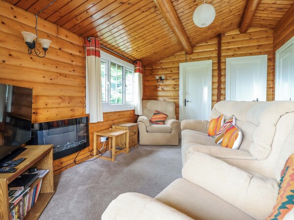 Interior de una cabaña de troncos con televisión, sofá, sillón y una pequeña mesa de madera.