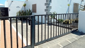 Une entrée fermée par un portail mène à un immeuble à la façade blanche, doté d’une antenne parabolique et agrémenté d’un petit jardin où poussent des cactus.