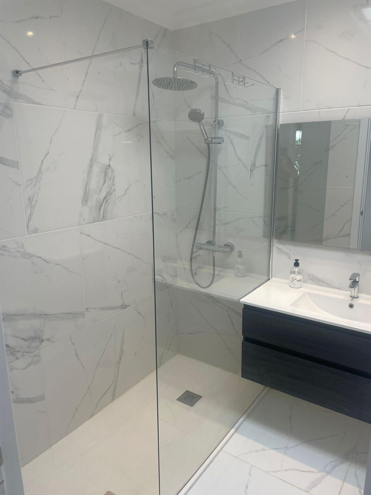 Une salle de bain moderne avec une douche à parois de verre, des carreaux de marbre, un lavabo doté d’un plan de travail foncé et un grand miroir.