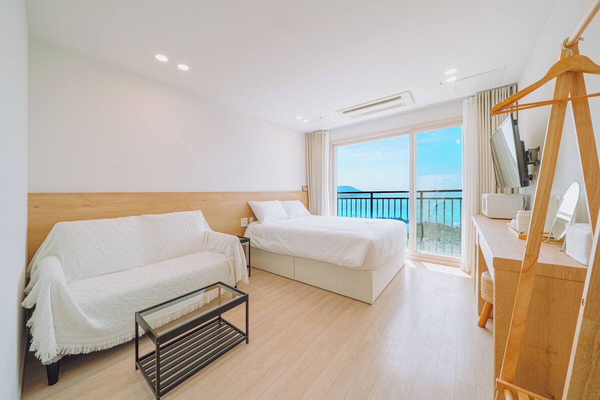 Une chambre d’hôtel moderne avec une tête de lit en bois, un canapé, un lit, un bureau avec un miroir et un balcon offrant une vue.