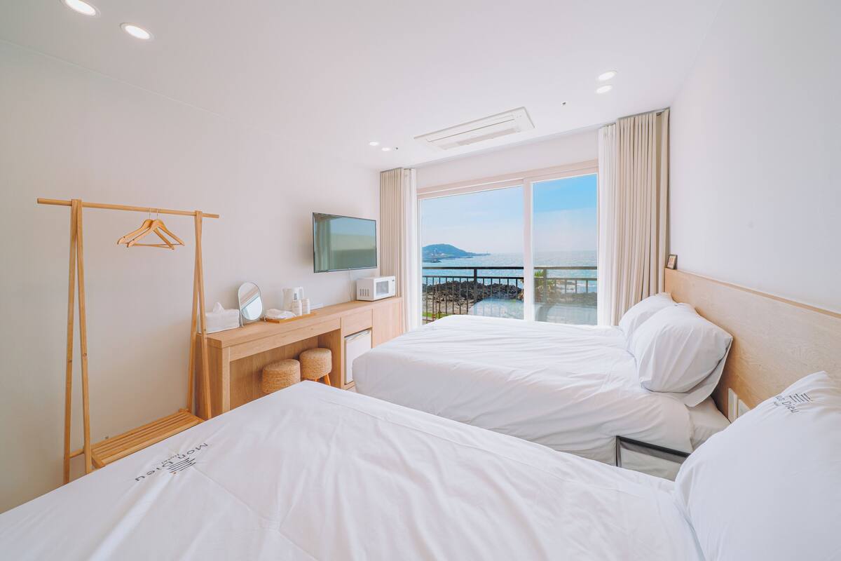 Une chambre d’hôtel avec deux lits, un bureau en bois avec une télévision, une fenêtre donnant sur la mer et un porte-manteau mural.