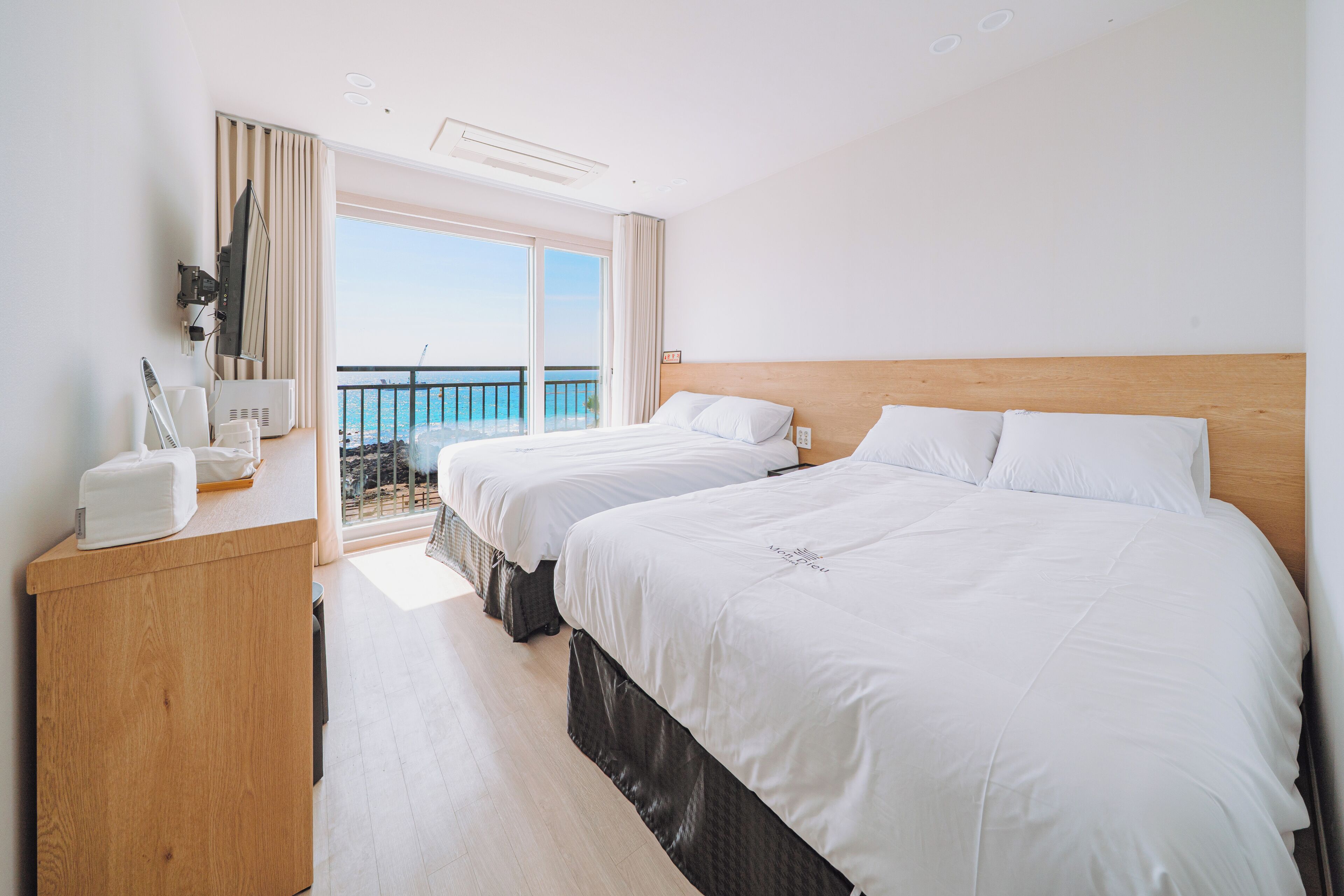 A hotel room with two beds, a wooden desk, a flat-screen TV, and a balcony with a view of the sea.