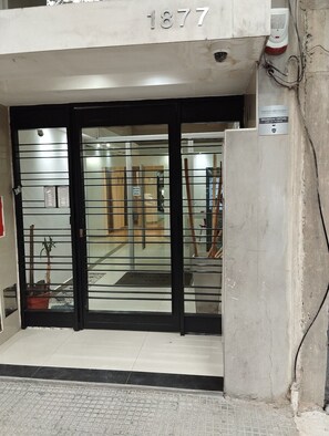 Une entrée d’immeuble avec une porte en verre, une plaque métallique portant un numéro et un panneau d’avertissement de sécurité.