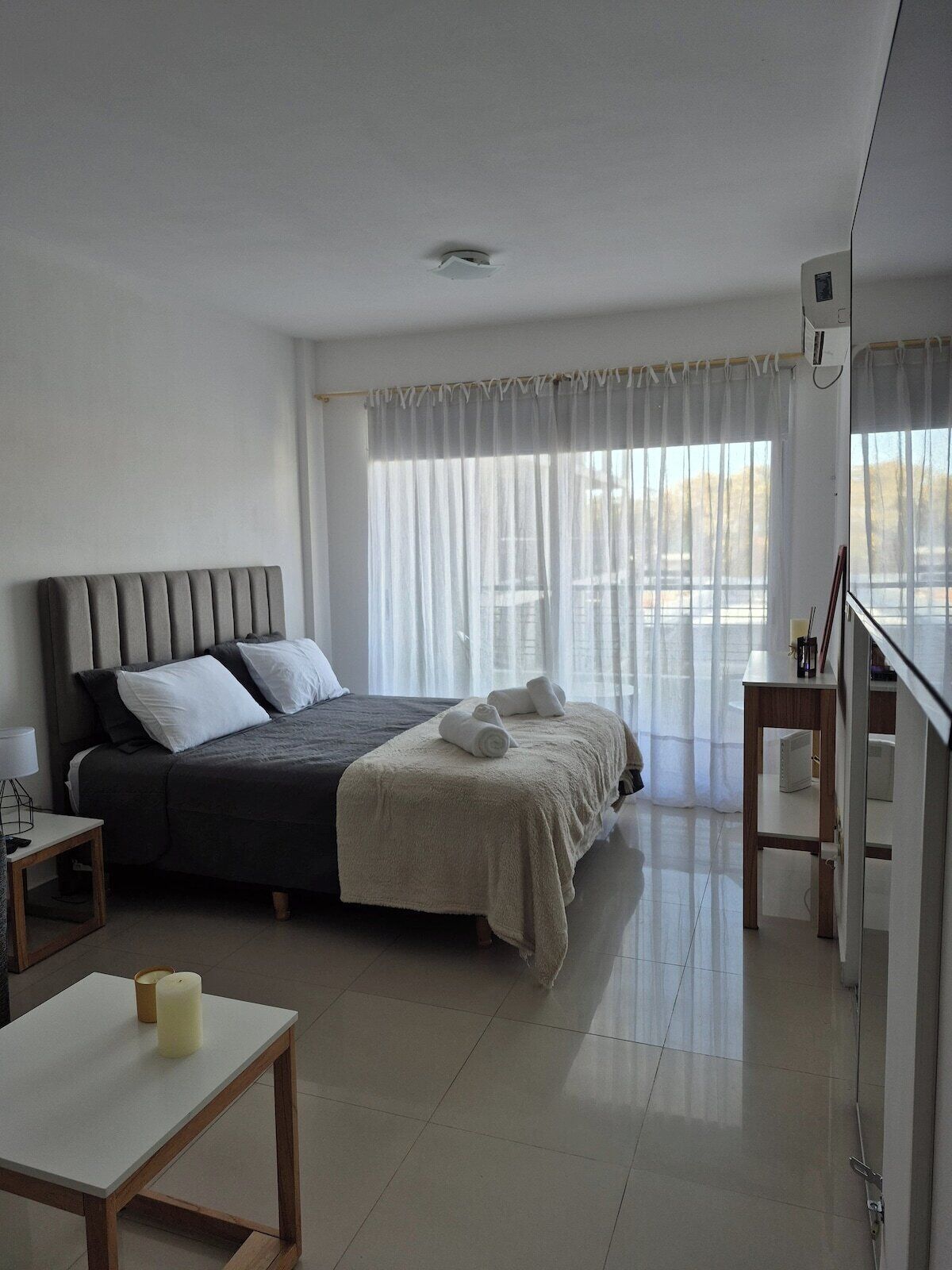 A hotel room with a bed, a bedside table with a lamp, a small table with a candle, and a window with sheer curtains.