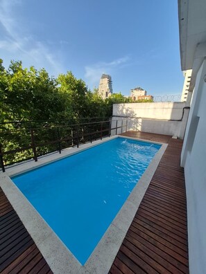 A rectangular pool with a wooden deck surrounding it, set against a backdrop of trees and buildings.