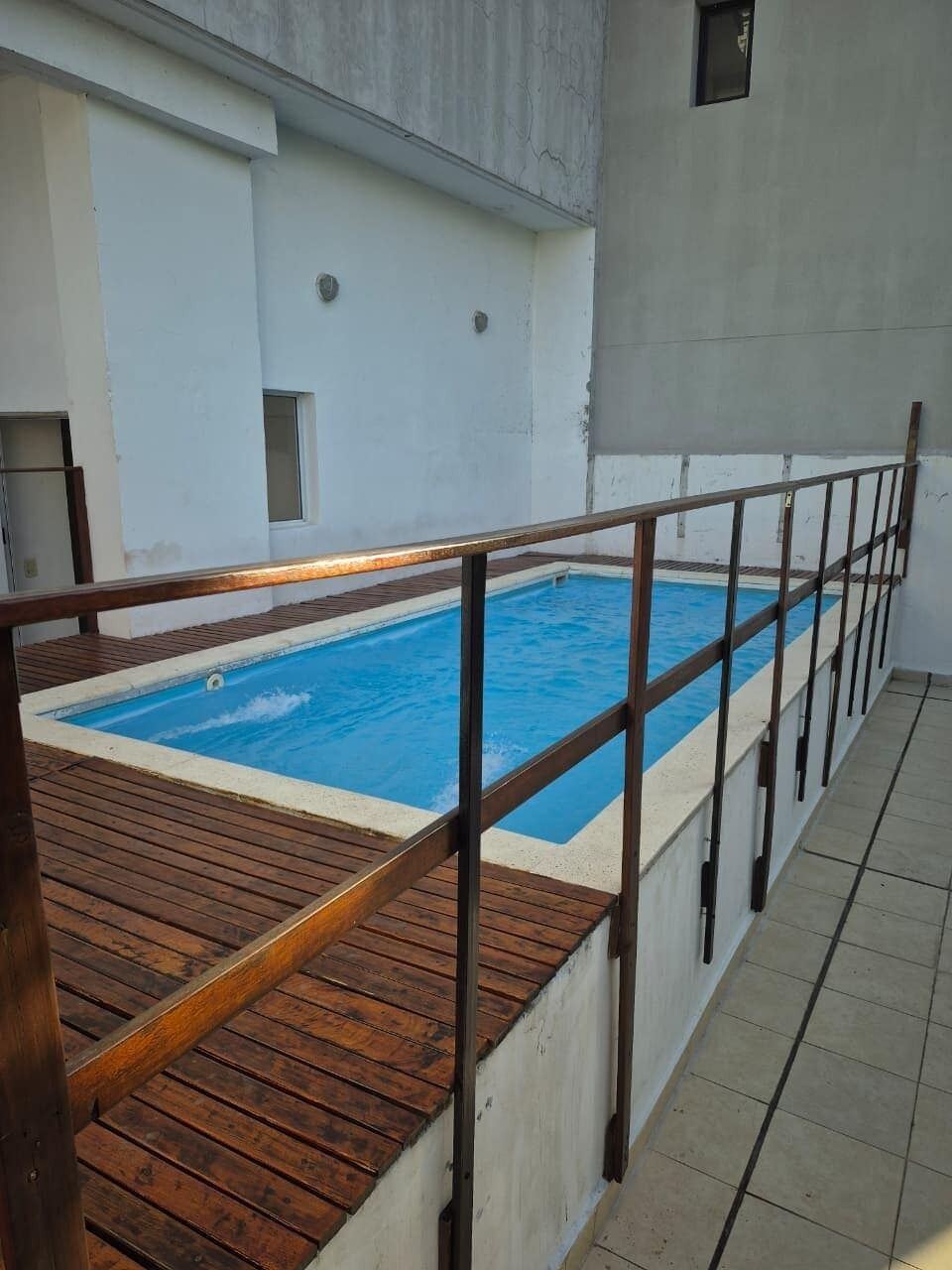 A rectangular swimming pool with a wooden deck and metal railing, surrounded by a white wall.