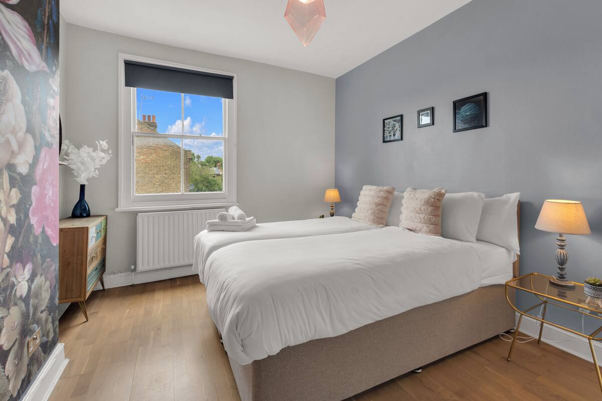 A bedroom with two beds, a window with a view, a radiator, and a bedside table with a lamp.