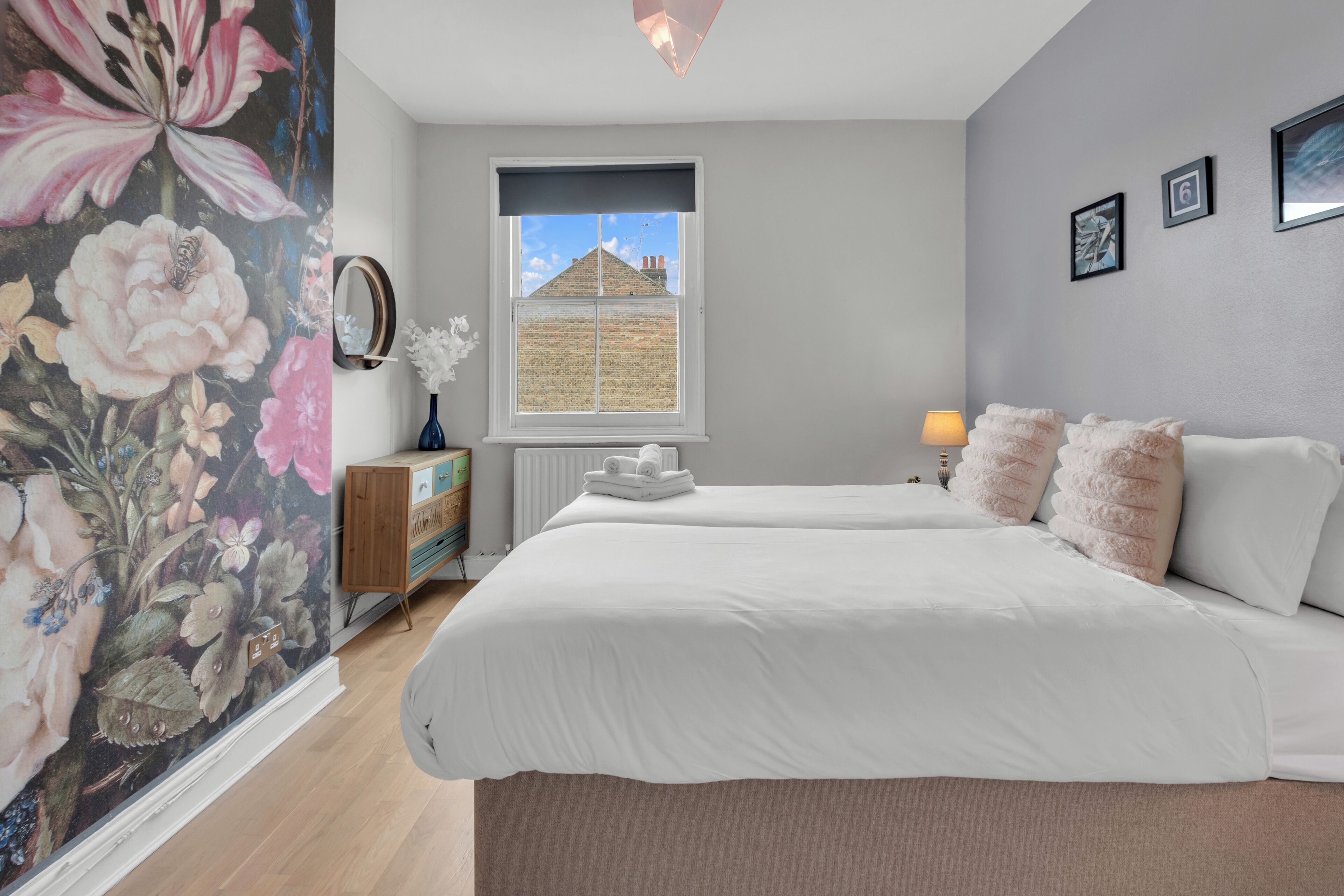 Une chambre avec deux lits, une fenêtre donnant sur des immeubles, un radiateur, une commode et une fresque florale sur le mur.