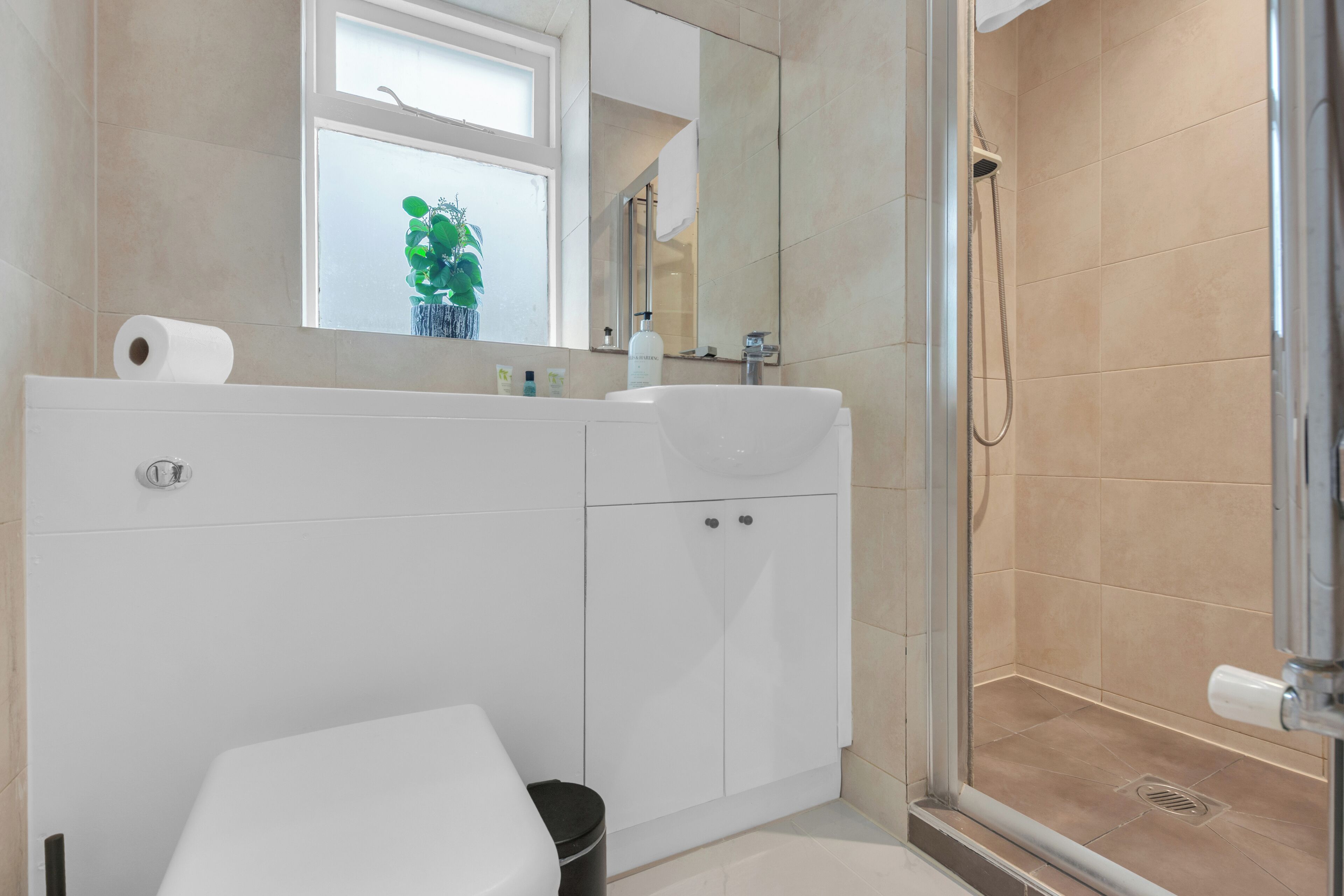 A modern bathroom with a white sink, mirror, and toilet. There is a shower area with a glass door and beige tiles.
