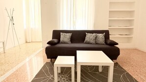 Una sala de estar con un sofá oscuro, dos mesitas blancas, una lámpara de pie y una ventana con cortinas traslúcidas.