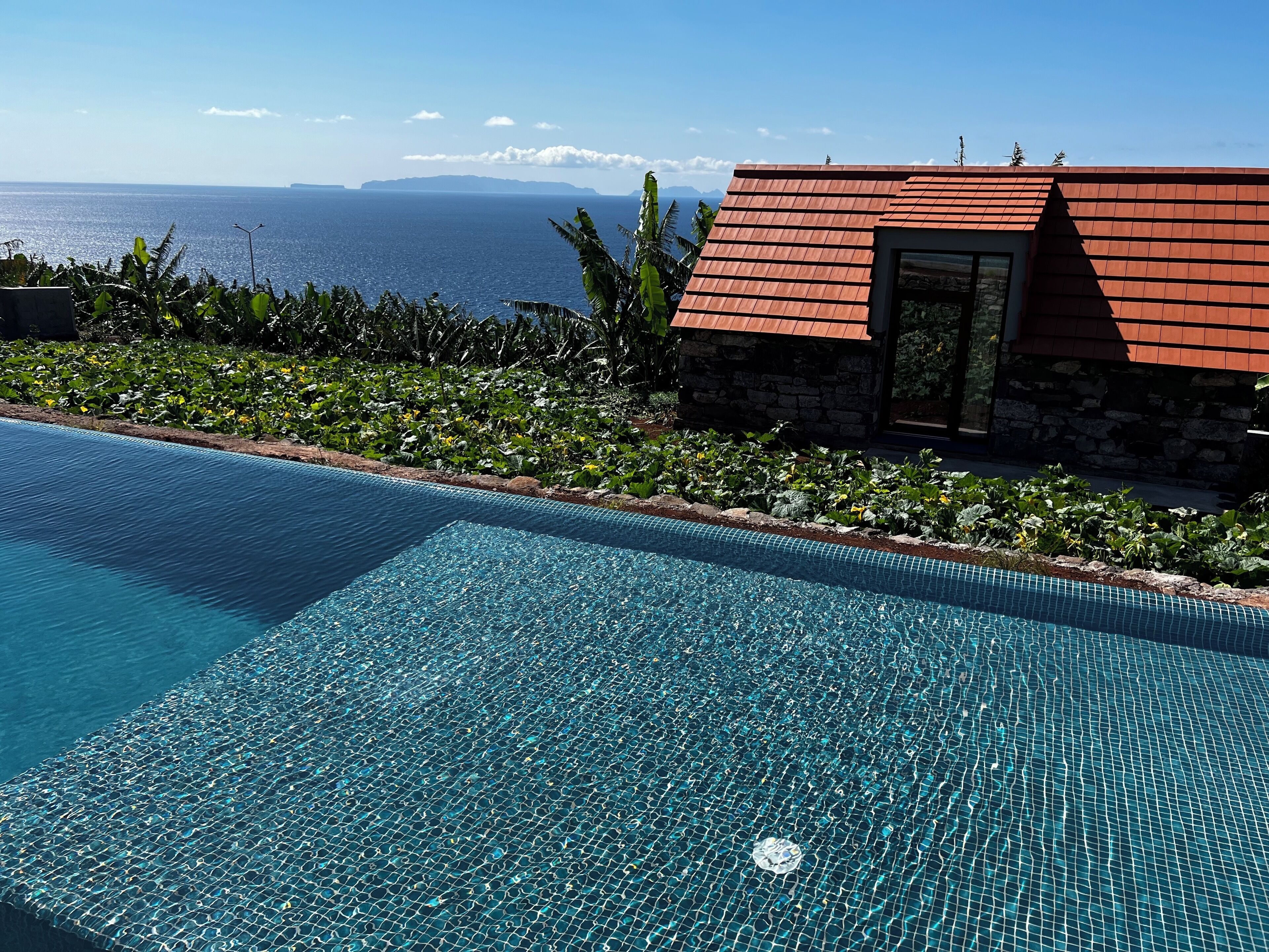 A clear blue pool with a tiled edge, a stone building with a red roof, and a view of the sea in the background.
