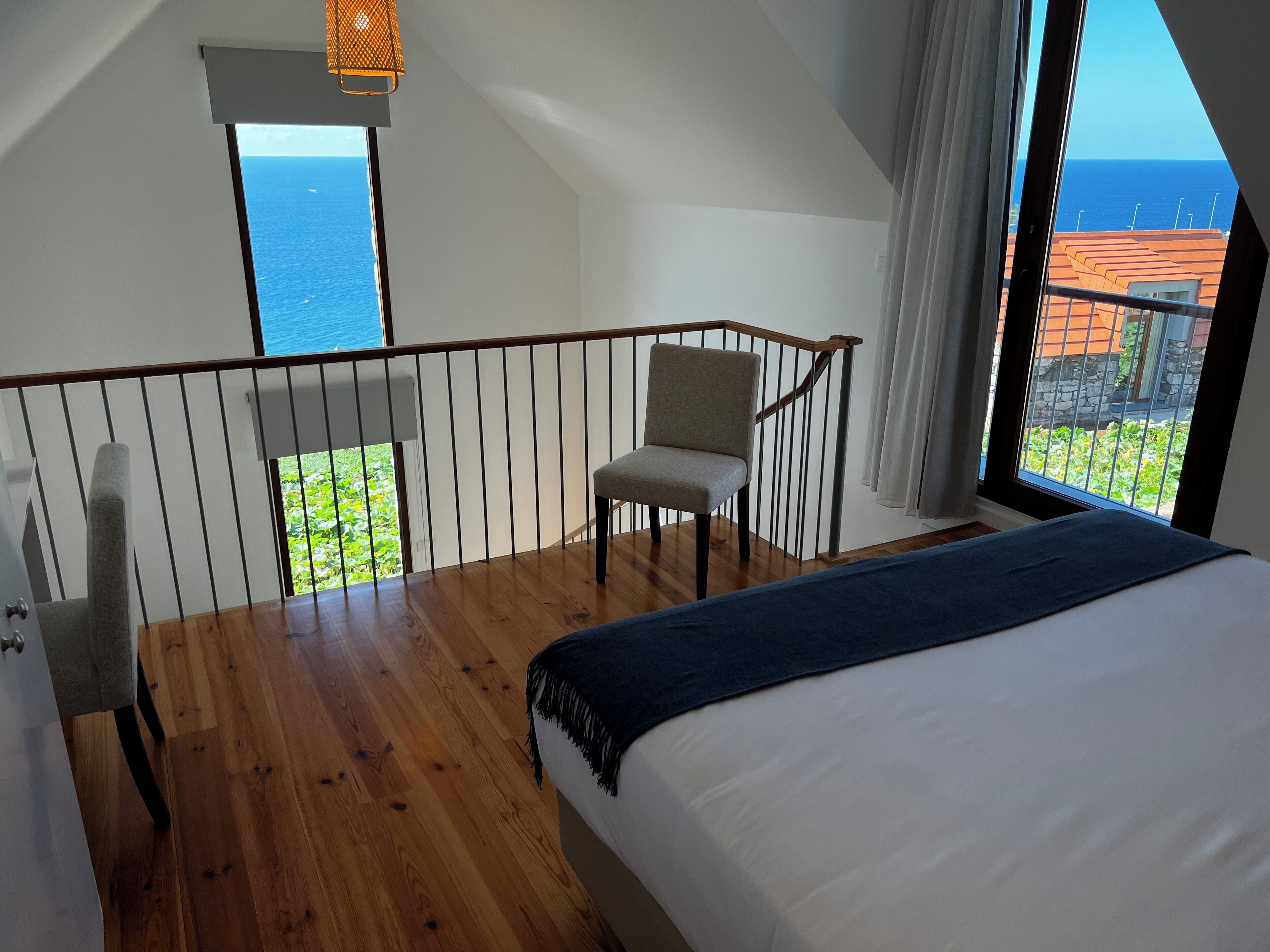 A bedroom with a large bed, a chair, a balcony with a view of the ocean, and a hanging light fixture.