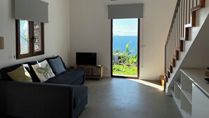 A modern living room with a sofa, a flat-screen TV, and a large window offering a view of the ocean. - CASAS DO LARANJO- Cottages Infinity Pool by brugrani (Santa Cruz)