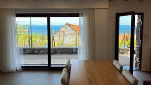 A modern room with a large wooden table, a stone wall, and a view of the ocean through large glass doors. - CASAS DO LARANJO- Cottages Infinity Pool by brugrani (Santa Cruz)