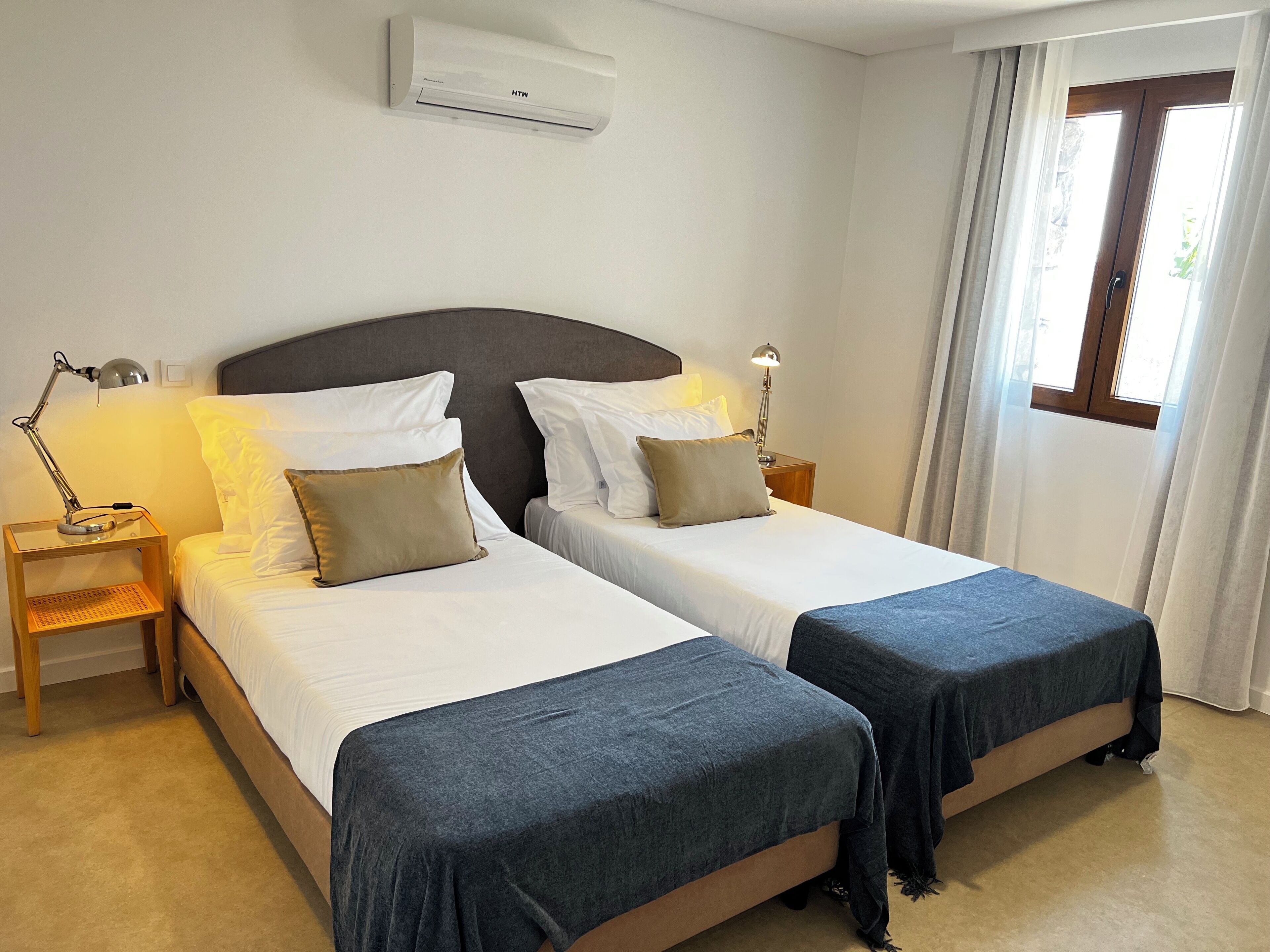 A hotel room with two single beds, a bedside table with a lamp, an air conditioning unit, and a window with curtains.
