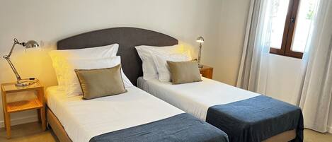 A hotel room with two single beds, a bedside table with a lamp, an air conditioning unit, and a window with curtains.