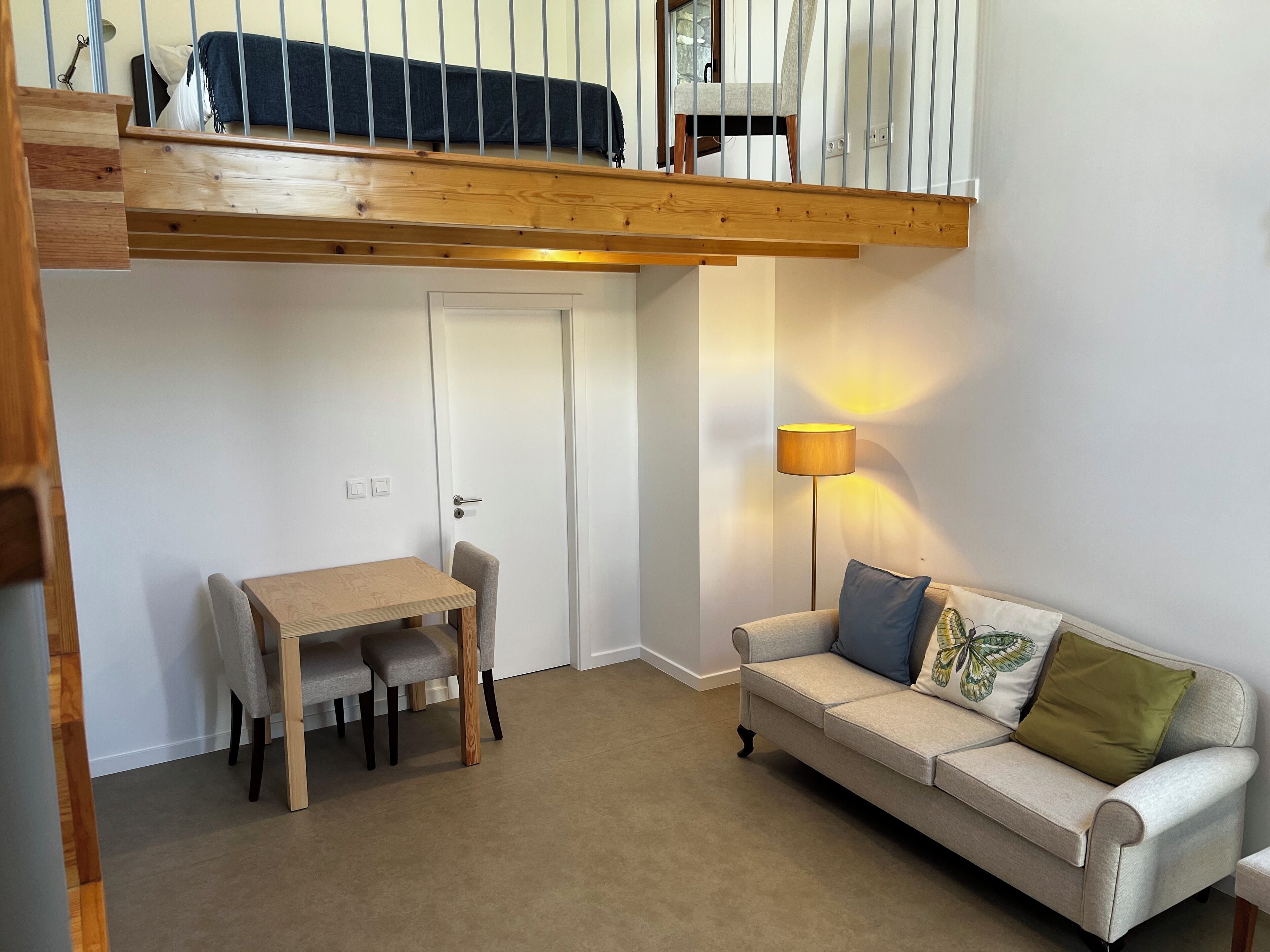 A loft-style bedroom with a wooden ceiling, a dining table with two chairs, a sofa with cushions, and a floor lamp.