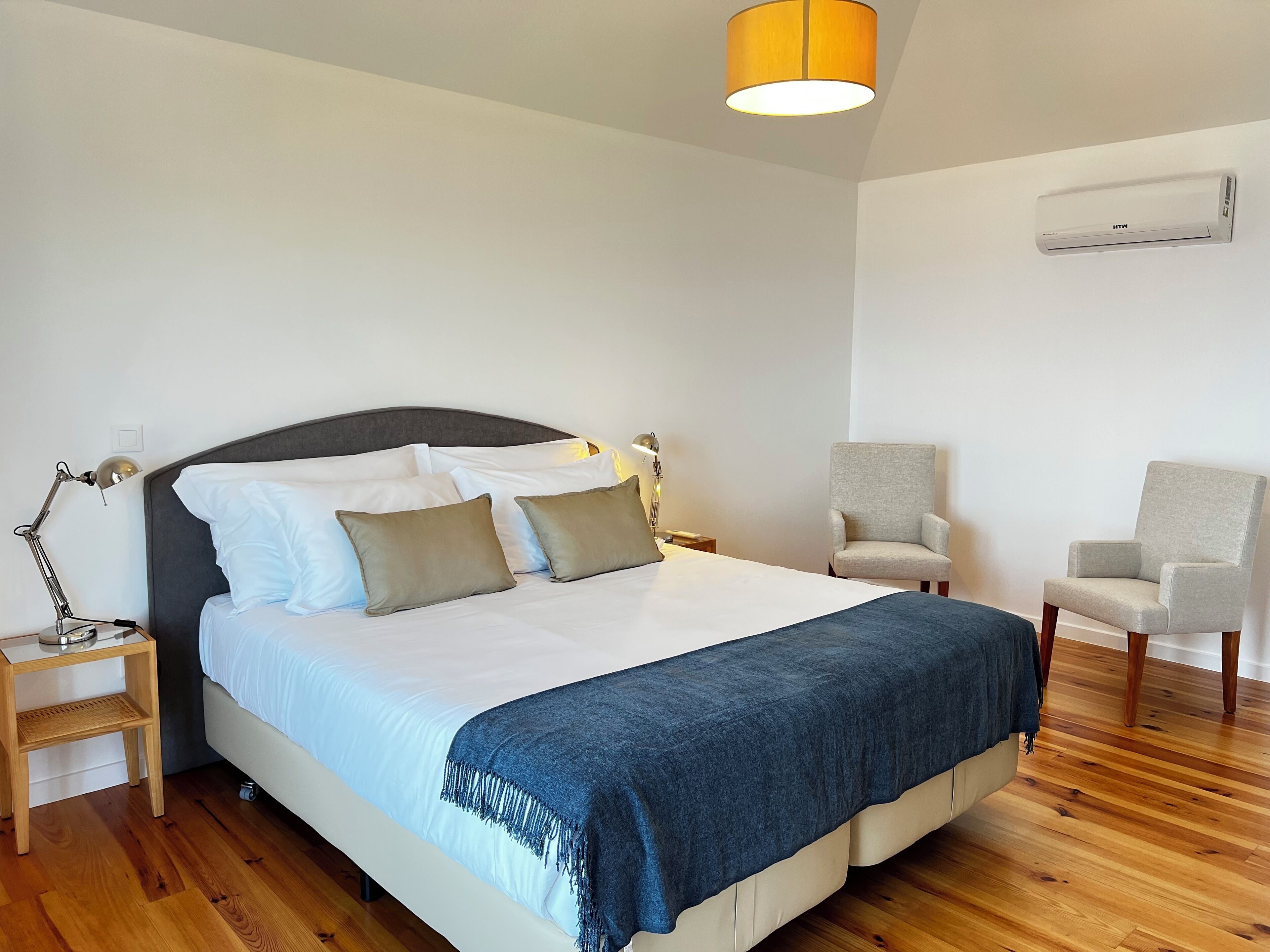 Un dormitorio ordenado con cama, lámparas de noche, una silla y un equipo de aire acondicionado en la pared.