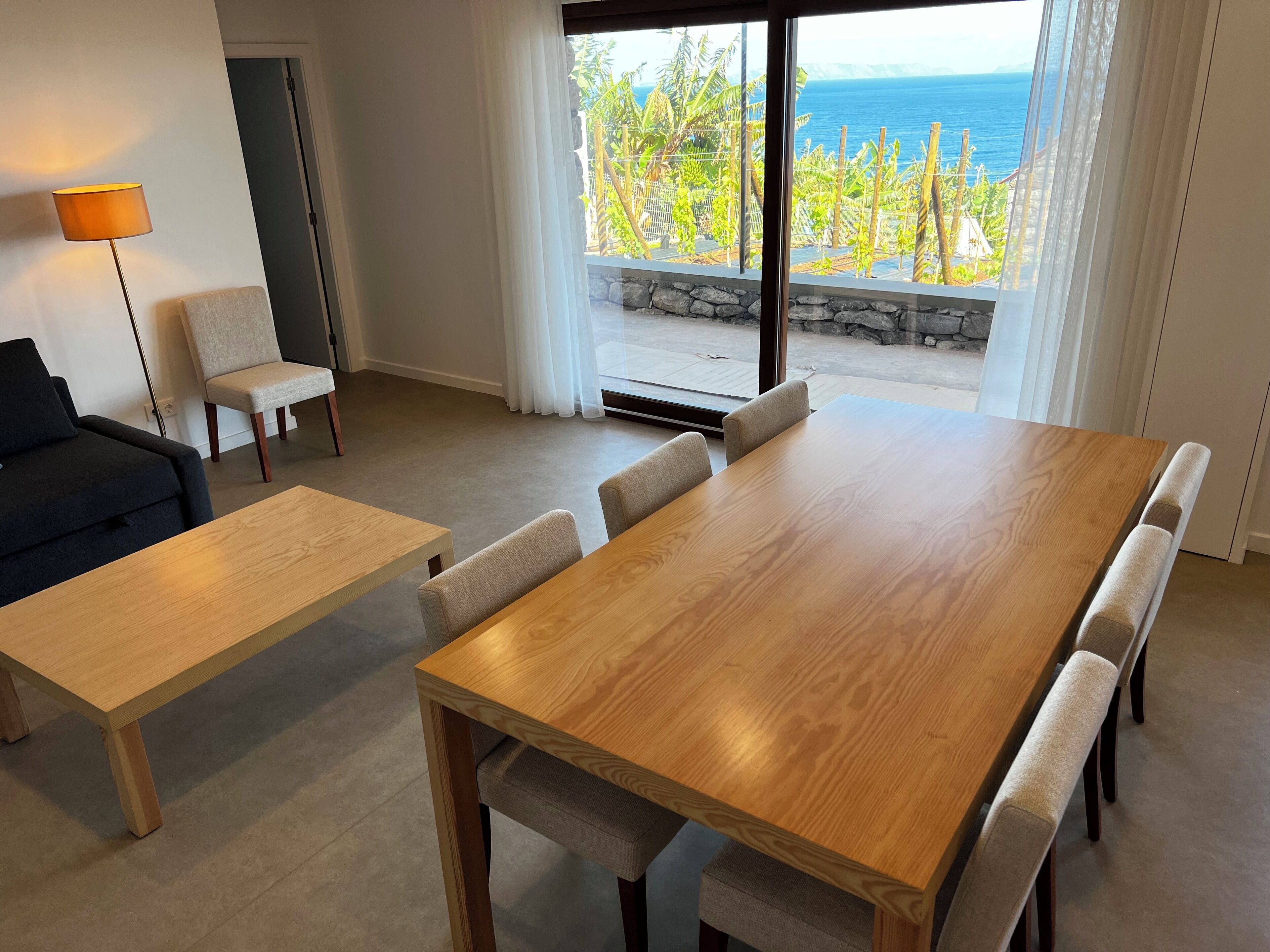 Een moderne woonkamer met een houten eettafel, een bank en een stoel. Een groot raam biedt uitzicht op de oceaan en groen.