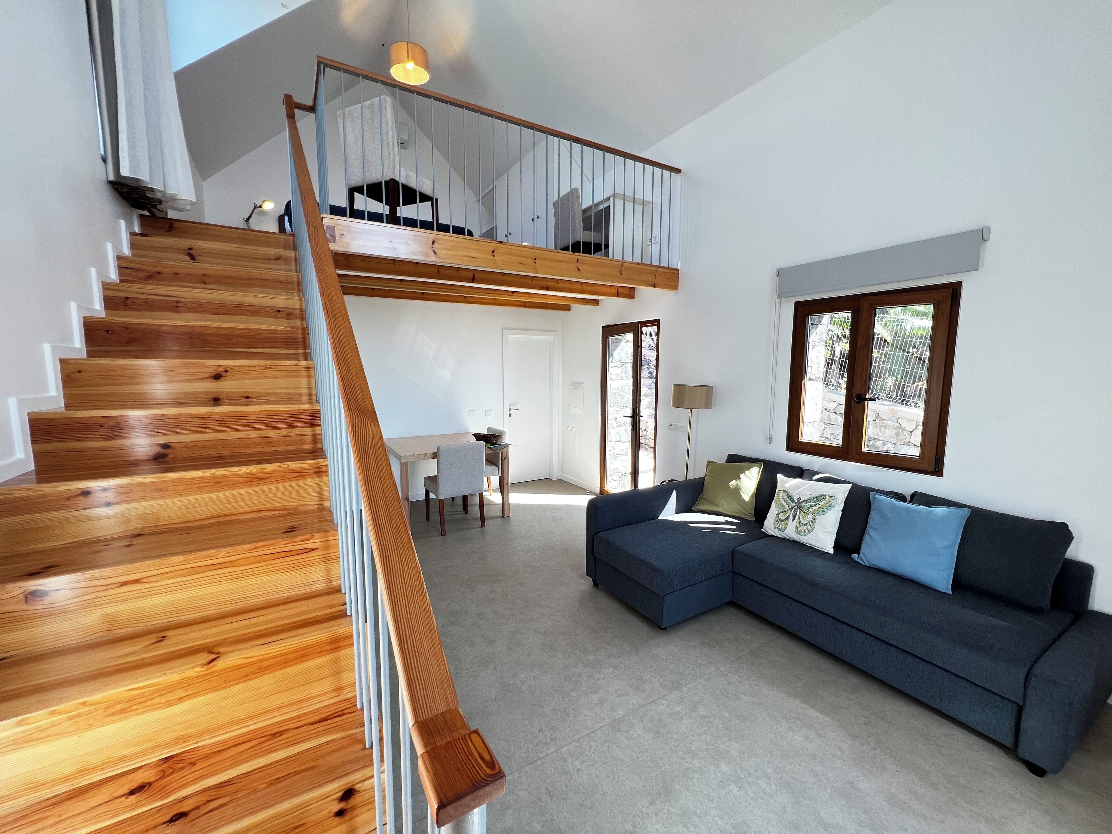 Un interior moderno con una escalera de madera que lleva a una habitación loft, un sofá azul oscuro y una zona de comedor con mesa y sillas.