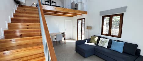 Un interior moderno con una escalera de madera que lleva a una habitación loft, un sofá azul oscuro y una zona de comedor con mesa y sillas.