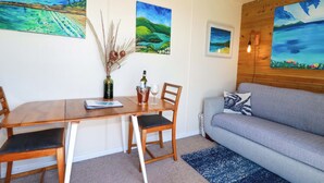 A dining area with a wooden table and chairs, a vase with dried plants, and a bottle of wine. There is a grey sofa with a patterned pillow in the living area. - The Studio (Truro)