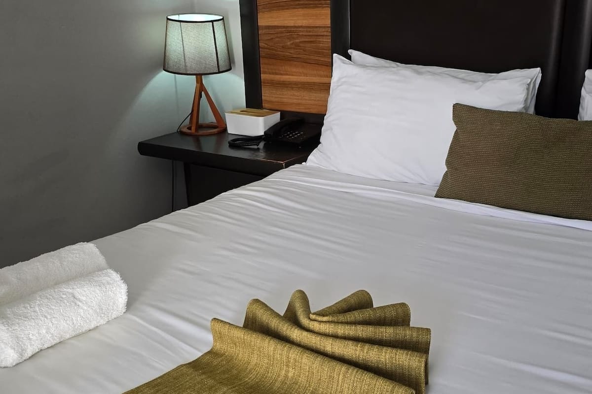 Una cama bien hecha con sábanas blancas, un edredón dorado y una almohada marrón. Junto a la cama, una mesita de noche con una lámpara y un teléfono.