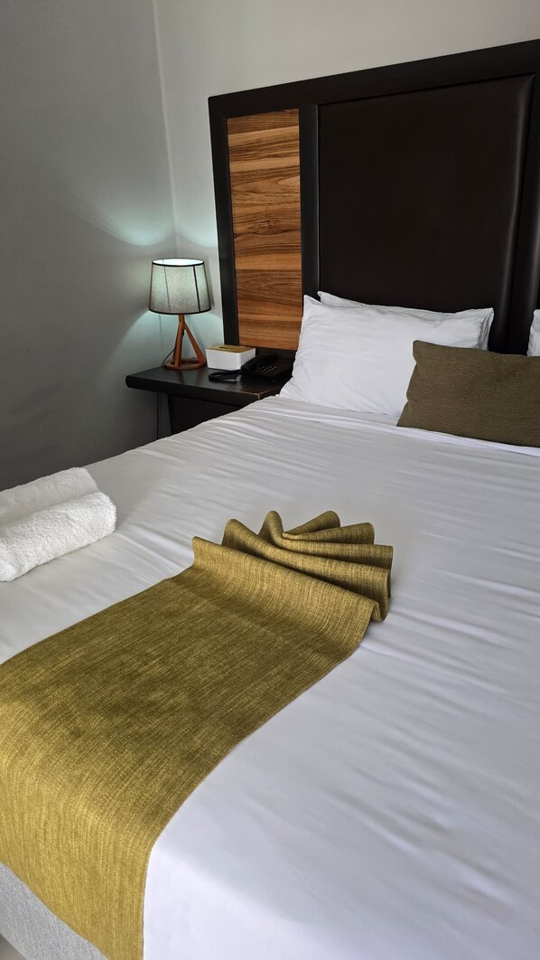 A neatly made bed with white linens, a gold throw blanket, and a brown pillow. There is a nightstand with a lamp and a telephone beside the bed.