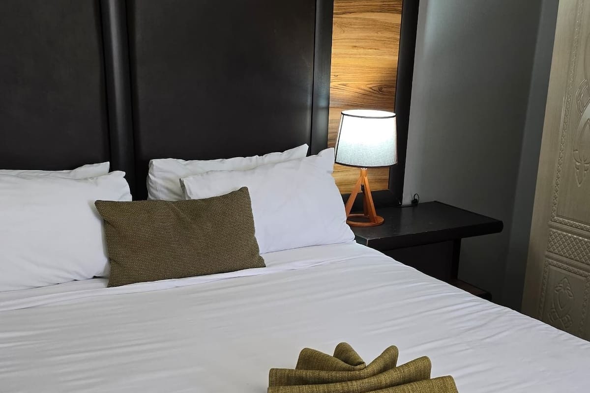 Habitación de hotel con una cama grande, dos almohadas, una mesita de noche con lámpara y una ventana con vistas.
