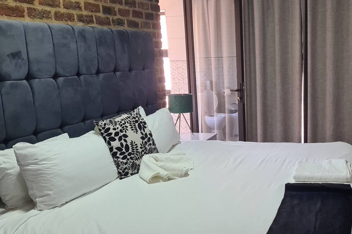 Habitación de hotel con una cama grande, cabecera, mesita de noche con lámpara, una silla de madera y vistas al exterior a través de una ventana con cortinas.