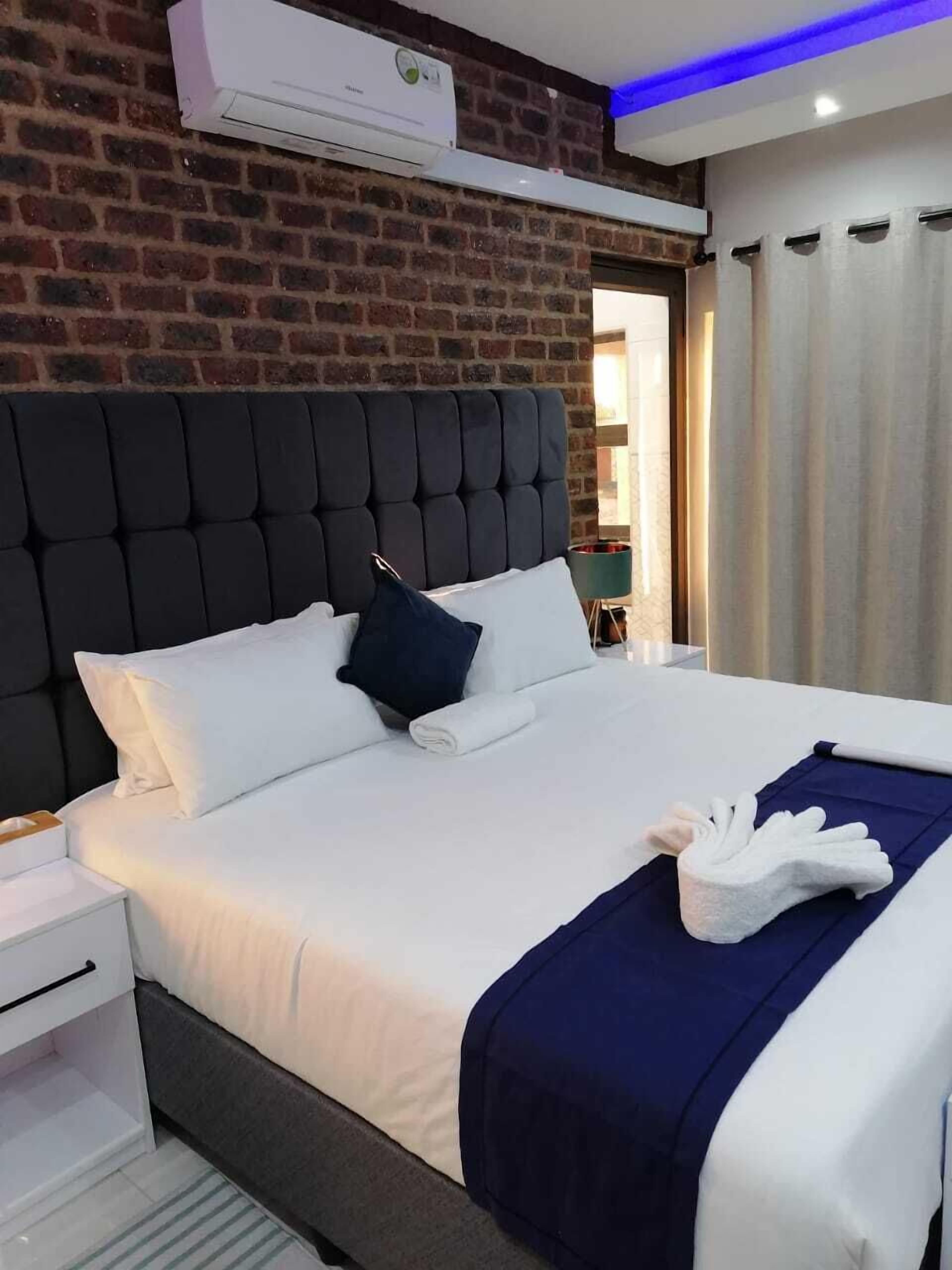 A modern hotel room with a brick wall, a tufted headboard, white bedding, and a blue accent pillow.
