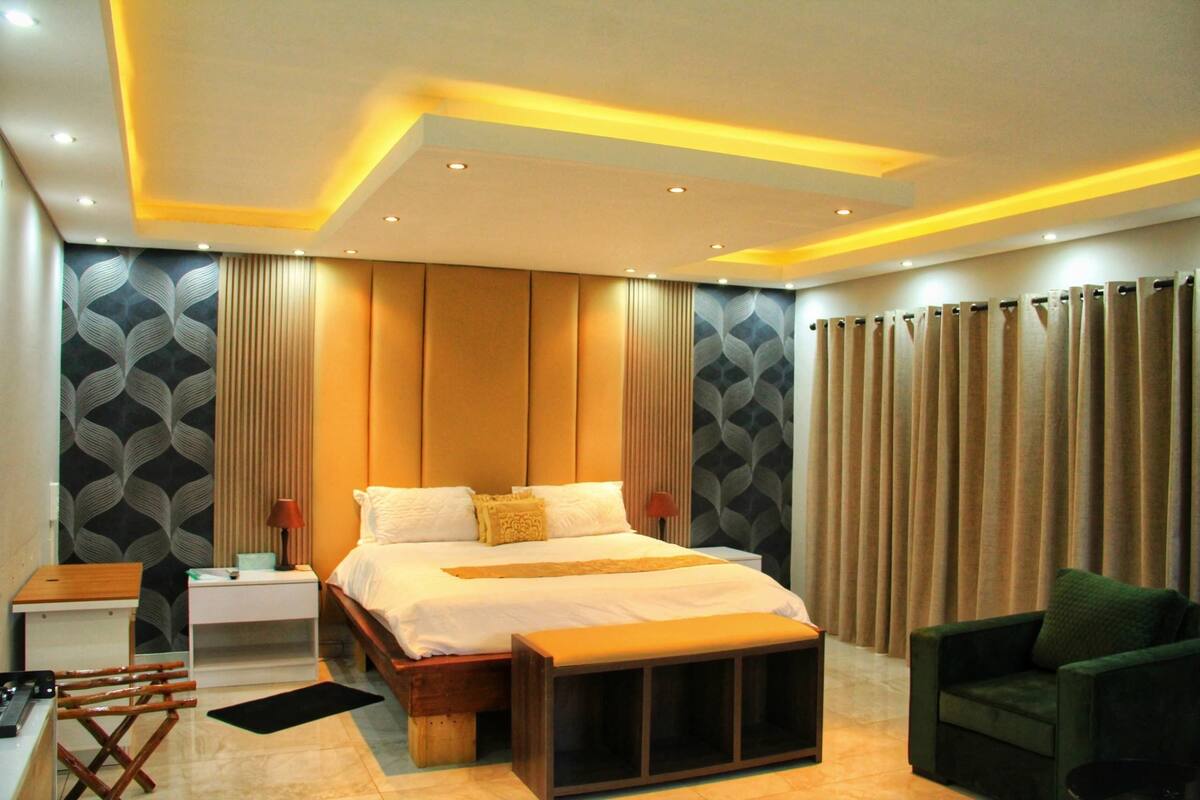 Una habitación de hotel moderna con una cama grande, un cabecero de madera, una mesita de noche con lámpara, un sillón verde y una ventana con cortinas.